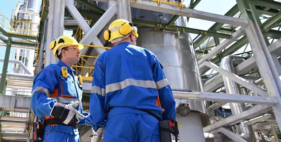 Two industrial workers wearing blue overalls and yellow safety helmets in a refinery; oil processing equipment and machinery in the background