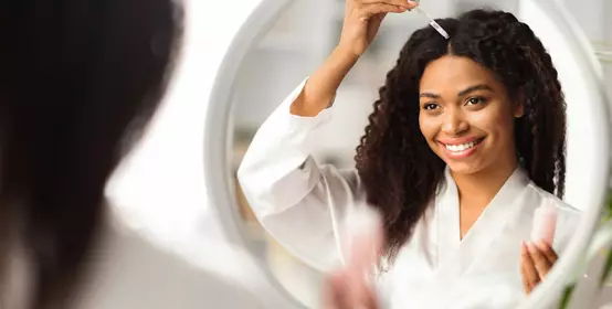 Smiling Black Woman Applying Serum For Hair Repair While Standing Near Mirror At Home, Young African American Female Making Haircare Treatment, Looking At Her Reflection, Selective Focus